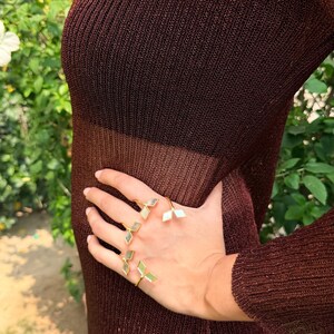 May include: A hand wearing several gold rings with geometric, light-colored inlays. The rings are displayed against a backdrop of a sheer, dark brown dress. The dress has a ribbed texture and is partially see-through.