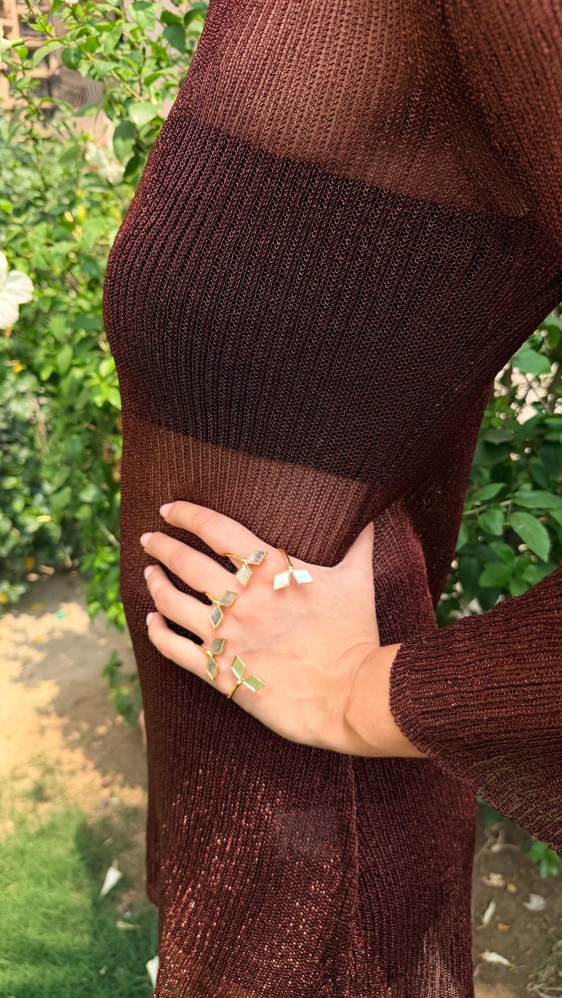 May include: A hand wearing several gold rings with geometric, light-colored inlays. The rings are displayed against a backdrop of a sheer, dark brown dress. The dress has a ribbed texture and is partially see-through.
