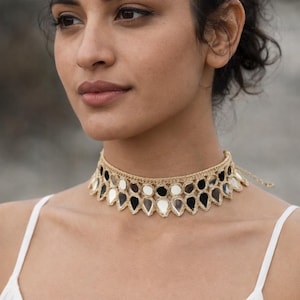 Op de afbeelding: Een goudkleurige choker-ketting met een herhalend patroon van druppelvormige spiegels. De spiegels wisselen af tussen zwart, wit en zilver. De ketting wordt om de nek gedragen.