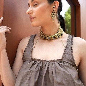 Può includere: Una donna indossa gioielli color oro, tra cui una collana girocollo e orecchini pendenti con dettagli neri. Gli orecchini sono collegati alla collana con una delicata catena. Indossa un top marrone.