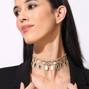 May include: A gold-toned choker necklace featuring a woven band with a series of dangling, oval-shaped reflective mirror accents. The necklace is worn around the neck and sits against a black blazer, showcasing a detailed metallic texture.
