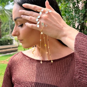 May include: A gold-toned hand chain and ring set with geometric white and clear accents. The jewelry features a delicate chain with dangling charms. The model is wearing a maroon-colored, long-sleeved top.