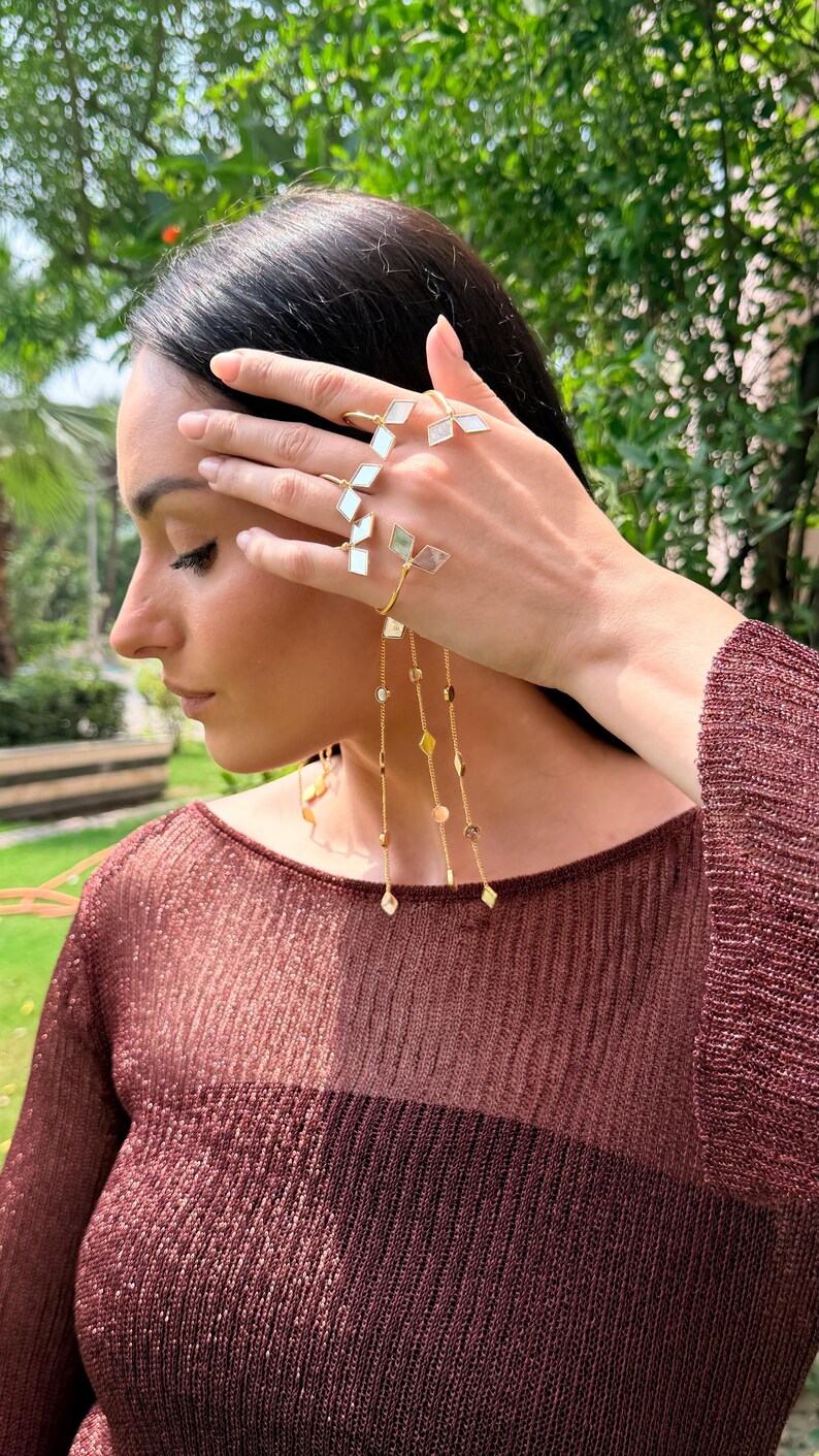 May include: A gold-toned hand chain and ring set with geometric white and clear accents. The jewelry features a delicate chain with dangling charms. The model is wearing a maroon-colored, long-sleeved top.