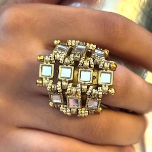 May include: Gold-coloured ring with a geometric design, featuring square mirrored insets. The ring has a textured appearance with small decorative elements. The design incorporates multiple layers and a symmetrical arrangement, creating a bold statement piece.