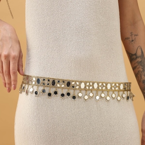 May include: A gold-coloured belt with small, reflective mirror accents and dangling embellishments. The belt is worn over a cream-coloured dress, creating a decorative waistline. The belt's design features geometric shapes.