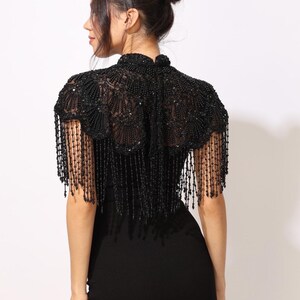Model wearing a black embroidered beaded cape with scalloped edges and fringe. The sheer net base is hand-embellished with intricate embroidery and sparkling black beads, creating a statement bridal or evening accessory.