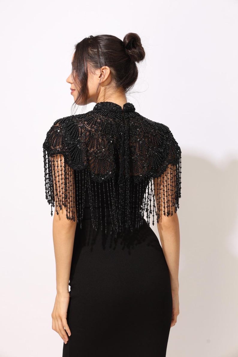 Model wearing a black embroidered beaded cape with scalloped edges and fringe. The sheer net base is hand-embellished with intricate embroidery and sparkling black beads, creating a statement bridal or evening accessory.