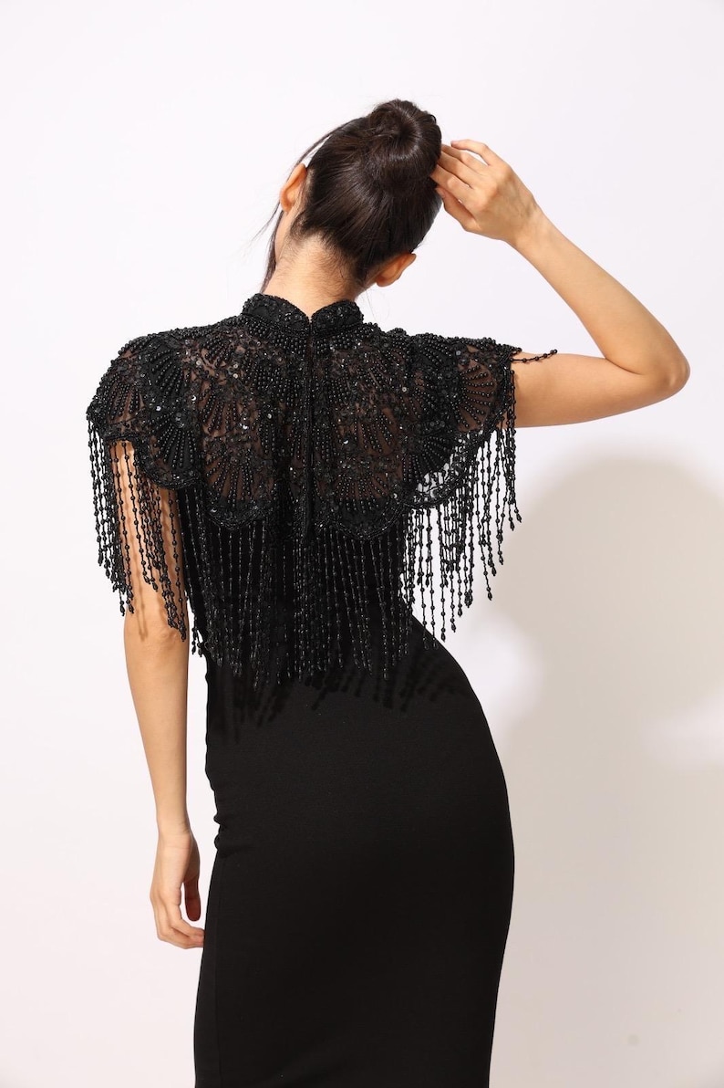 Model wearing a black embroidered beaded cape with scalloped edges and fringe. The sheer net base is hand-embellished with intricate embroidery and sparkling black beads, creating a statement bridal or evening accessory.