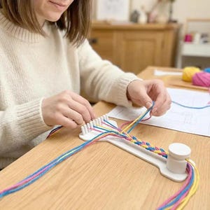 以下が含まれることがあります： 一端に一連のペグ、もう一端に円筒形の支柱を備えた白いプラスチック製の編み込みツール。このツールを使用して、鮮やかな黄色、ピンク、青の毛糸を複数本編み込み、平らな多色の組紐を作成しています。