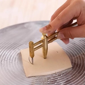 May include: A brass pottery circle cutter tool with an adjustable arm and a small metal blade, positioned to cut a circular shape into a square piece of light-colored clay resting on a circular, grooved metal pottery wheel surface.