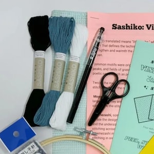 以下が含まれることがあります： 黒、青、白の刺繍糸3束、黒いマーキングペン、黒いハサミ、木製の刺繍枠、青い針ケース、そして「Sashiko: Visible Mending」というテキストが書かれた説明書が入った刺し子刺繍キット。