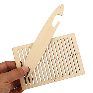 May include: A light-coloured wooden weaving loom tool set consisting of a rectangular board with vertical slots and circular holes, paired with a long, slender wooden shuttle featuring a notched end and a small hole at the pointed tip.