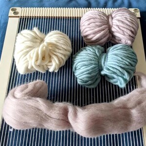 Può includere: Un telaio rettangolare in legno con fili di ordito bianchi verticali, che presenta tre gomitoli di filato spesso color crema, rosa antico e verde salvia, oltre a una lunga ciocca di morbida fibra pettinata rosa chiaro posata sul fondo.