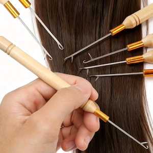 May include: A set of hair ventilation needles featuring light-coloured wooden handles with gold-toned metal ferrules and thin steel hooks. The tools are arranged over a background of straight, dark brown hair, with one needle held in a hand.