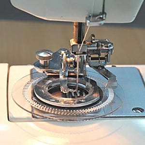 May include: A circular sewing machine attachment featuring a clear plastic disc with a serrated outer edge, a central metal ring with a plus and minus sign, and a silver metal presser foot assembly with a needle and a small adjustment screw.