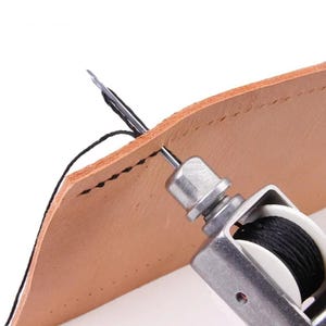 Op de afbeelding: Een metalen lederen stikgereedschap met een zilveren afwerking en een witte spoel met zwarte draad. De naald is door een stuk beige leer gestoken en trekt een zwarte draad door een voorgeponst gat om een zichtbare stiklijn te creëren.