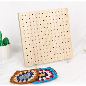 May include: A light-colored wooden pegboard with numerous small holes, supported by clear plastic stands. Two colorful, crocheted squares in shades of brown, blue, and red are in front of the board. A small cactus in a white pot is visible on the left.