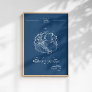 May include: Framed blueprint of a snare drum, with detailed technical drawings in white against a deep blue background. The print includes diagrams and text, showcasing the drum's components and design. The frame is a light wood color.