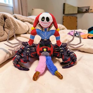 Può includere: Un peluche raffigurante una figura simile a un clown con un viso bianco, capelli rossi e un abito blu e rosso. Il giocattolo ha un corpo simile a un granchio con più zampe e un design colorato. Il peluche è su un letto.
