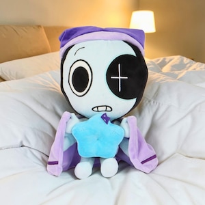 May include: A plush doll with a light blue face, black eye patch, and a purple nightcap. The doll has a blue star-shaped body and purple cape-like arms. The doll is sitting on a white bed.