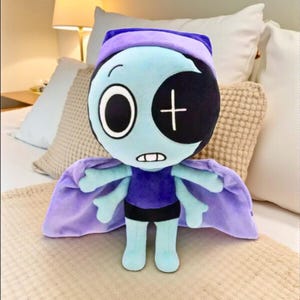 May include: A plush doll with a cartoonish design. The doll has a light blue face with a black eye patch and a white cross. It wears a purple outfit and cape. The doll is standing on a bed.