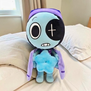 May include: A plush toy in the shape of a cartoon character with a light blue face, a black eye patch, and a white cross. It has a purple hood and cape, and a blue star-shaped body. The toy is on a white bed.