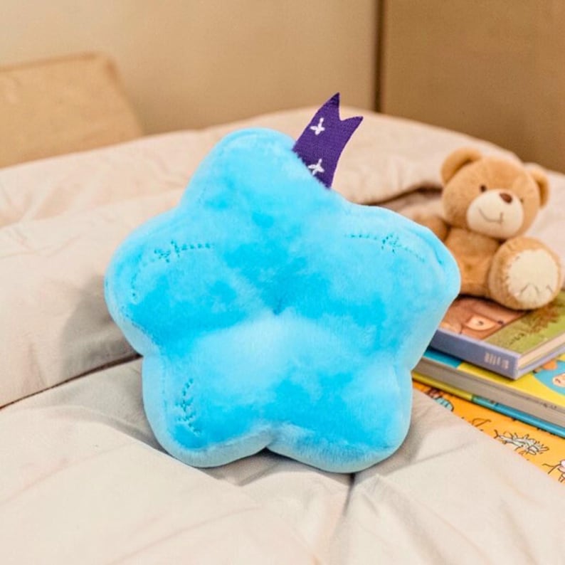 May include: A plush, light blue star-shaped pillow with a purple accent. The pillow is on a white bedspread, with a teddy bear and books in the background. The star has a stitched outline and a soft, fluffy texture.