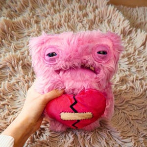 Può includere: Un giocattolo a forma di mostro di peluche rosa con un'espressione triste, che tiene in mano un cuore rosso con una benda cucita. Il mostro ha grandi occhi cuciti e una bocca larga. Il giocattolo è fatto di materiale morbido e sfocato.