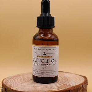 May include: A brown glass bottle of cuticle oil with a black dropper, labeled "Nature's Gold Cuticle Oil." The label includes the text "You're Kinda Cute" and "1 oz." The bottle sits on a wooden slice.