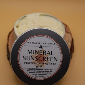 May include: A round, open tin of mineral sunscreen with a black lid, sitting on a wooden slice. The lid features the text "MINERAL SUNSCREEN" and "PROTECT & HYDRATE". The product contains 2 oz of sunscreen.