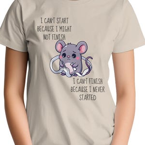 May include: A beige t-shirt featuring a graphic of a small gray mouse holding a long white paper scroll. The text reads: I CAN'T START BECAUSE I MIGHT NOT FINISH and I CAN'T FINISH BECAUSE I NEVER STARTED. The mouse has large ears and a sad expression.