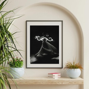 Könnte beinhalten: Schwarz-weiß gerahmtes Kunstwerk einer Person in einem fließenden Kleid, das von einem leuchtenden, wirbelnden Licht beleuchtet wird. Das Kunstwerk wird auf einem Holzregal mit einer Pflanze in einem weißen Topf und Büchern ausgestellt.
