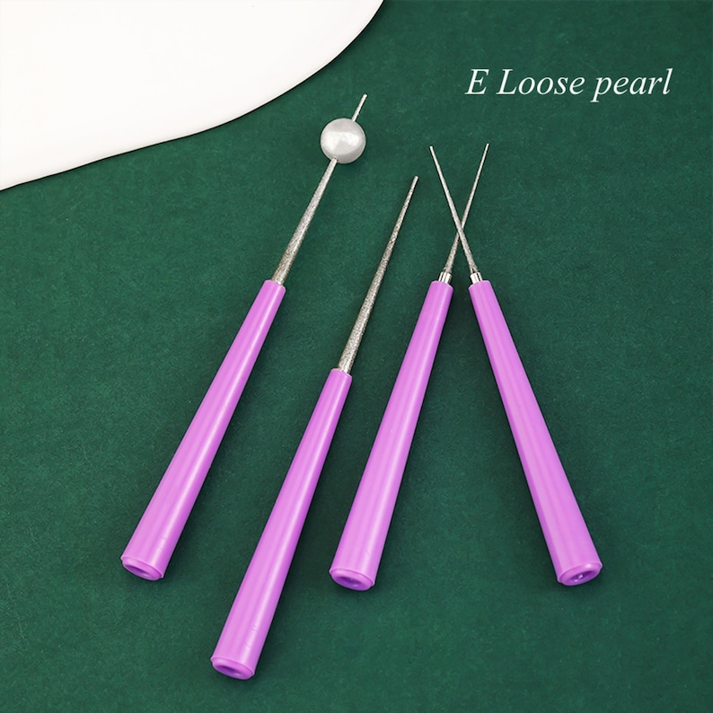 May include: Three purple tools for threading pearls. The tools have a pointed tip and a handle. The tools are on a green surface. E Loose pearl.