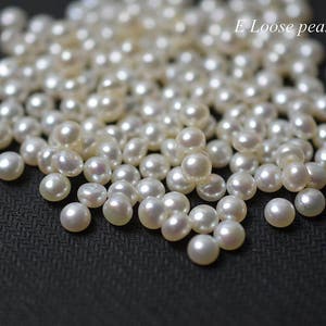 Puede incluir: Primer plano de un montón de perlas blancas sueltas. Las perlas son redondas y lisas, y están esparcidas sobre una superficie negra. E Loose pearl está escrito en texto blanco en la esquina superior derecha de la imagen.