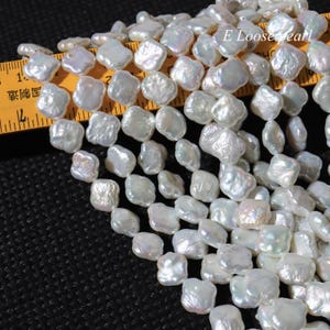 Może przedstawiać: Stos nieregularnych, białych pereł barokowych. Perły mają opalizujący połysk. Żółta linijka z czarnymi oznaczeniami jest częściowo widoczna. Perły są rozrzucone na czarnej powierzchni. Widoczny jest tekst "E Loose pearl".