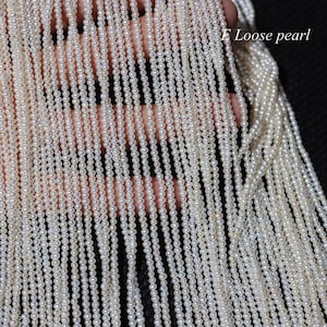 Op de afbeelding: Een streng witte zoetwaterparels, geregen tot een lange lijn. De parels zijn klein en uniform van grootte, en ze hebben een gladde, glanzende afwerking. De streng wordt in een hand gehouden, en de achtergrond is zwart.