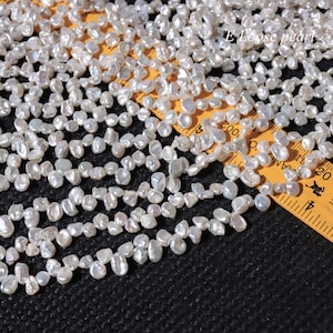 Puede incluir: Primer plano de una colección de perlas sueltas blancas e irregulares. Las perlas están dispersas sobre una superficie oscura, con una regla amarilla parcialmente visible. El texto "E Loose pearl" está en la esquina superior derecha.