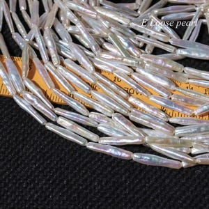以下が含まれることがあります： 白く虹色の光沢のある、ばら売りのライス型の真珠の糸。真珠の長さは約2.5cmで、定規の上に並べてあります。E Loose pearl.