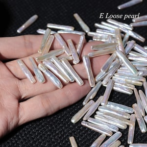 May include: A close-up of a hand holding a pile of white, iridescent, long, thin, pearl beads. The beads are scattered on a black surface. The text "E Loose pearl" is visible in the upper right corner of the image.