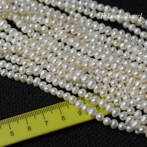 Puede incluir: Una hebra de perlas de agua dulce blancas, que mide aproximadamente 23 cm de largo, se muestra en una regla amarilla. Las perlas son redondas y lisas, con una ligera variación de tamaño. E Loose pearl.