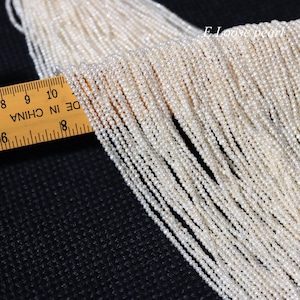 Può includere: Primo piano di una collana di perle d'acqua dolce bianche, misurata con un righello in pollici. Le perle sono piccole e di dimensioni uniformi, creando un aspetto delicato ed elegante.