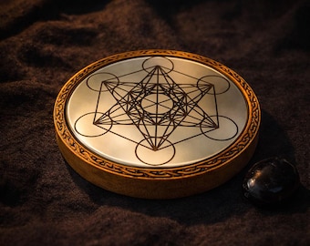 May include: A round, wooden coaster with a metallic surface featuring a Metatron's Cube design. The coaster has a decorative border and sits on a dark, textured surface. A small, black stone is next to the coaster.