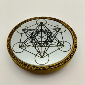 May include: Round wooden coaster with a mirrored surface featuring a black Metatron's Cube design. The coaster has a gold-colored border with an intricate carved pattern. The design is centered on the reflective surface.