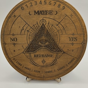 Op de afbeelding: Een rond houten waarzegbord met geëtste ontwerpen en tekst. Het bord bevat de woorden "MAYBE", "YES", "NO" en "REFRAME". Cijfers en symbolen zijn langs de randen geëtst, met een oog-in-driehoek-symbool in het midden. Het bord is lichtbruin.