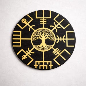 Pode incluir: Uma decoração de parede redonda de madeira preta com um desenho dourado da árvore da vida no centro, rodeada por intrincados símbolos geométricos. A obra de arte tem um aspeto rústico e envelhecido.