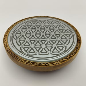 Op de afbeelding: Een ronde houten onderzetter met een glazen bovenkant met een grijs Flower of Life-patroon. De onderzetter heeft een lichtbruine, gegraveerde houten rand. De onderzetter heeft een diameter van ongeveer 10 cm.