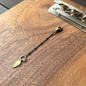 May include: A gold-colored leaf charm attached to a dark brown beaded chain. The chain has small black beads and a gold clasp. The charm is a detailed feather shape. The background is a wooden surface with a silver metal object.