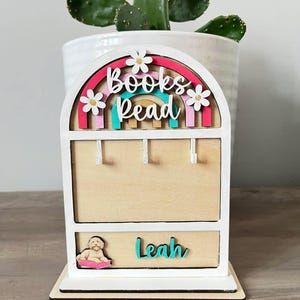 Peut inclure: Étagère en bois avec un design en arc-en-ciel, les mots "Books Read" sont affichés en lettres colorées. L'étagère a des crochets pour suspendre des objets et un tiroir avec le nom "Leah". Une petite illustration de chien est sur le tiroir.