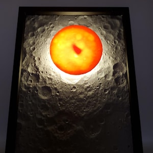 May include: A framed, illuminated artwork featuring a textured, gray surface resembling the moon's surface. A bright orange, circular light source is positioned centrally, mimicking a planet. The artwork is encased in a black frame, with a white power cord extending from the bottom.