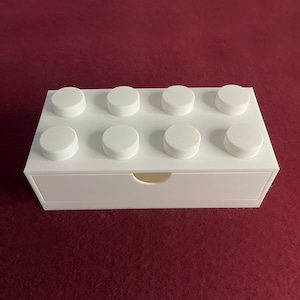 Puede incluir: Una caja de almacenamiento blanca diseñada para parecerse a un gran bloque de construcción. La caja tiene ocho clavijas circulares en la parte superior y un cajón con un asa curva. La caja está hecha de un material blanco sólido.